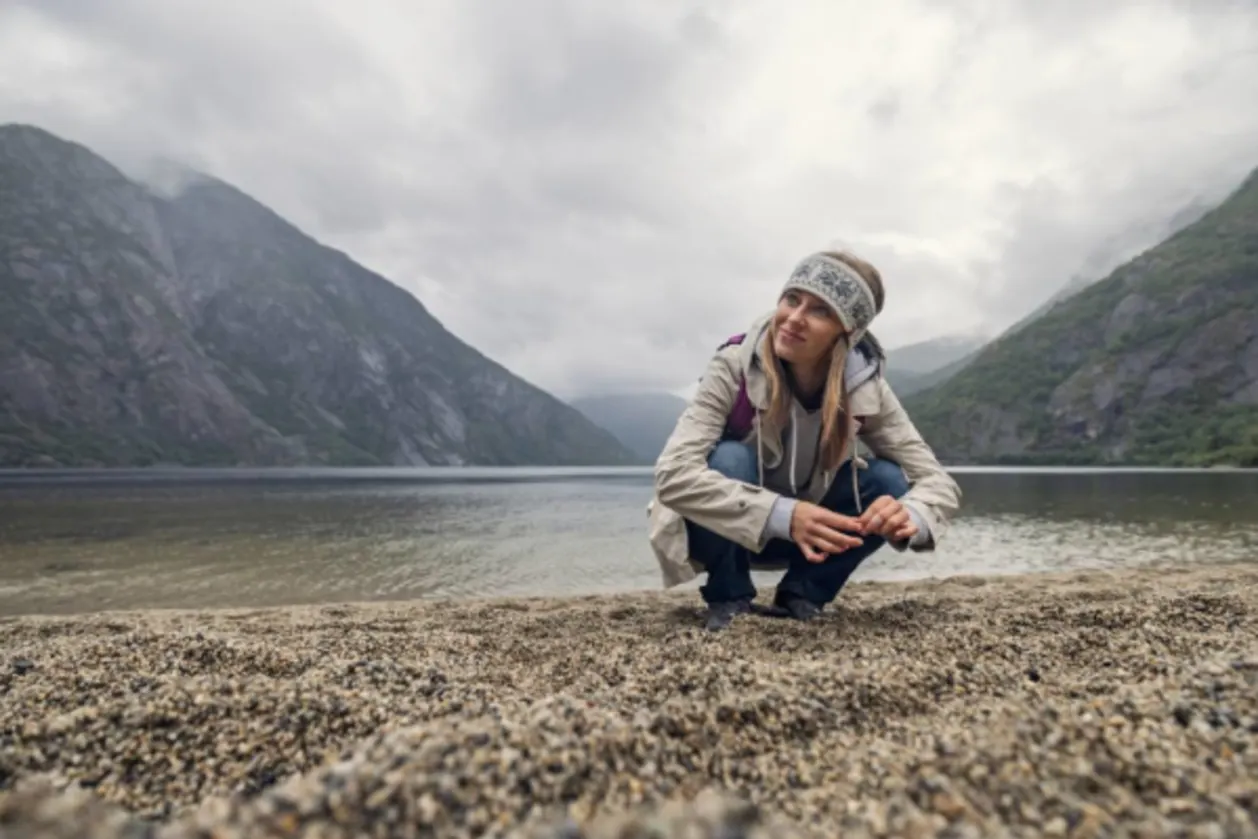 Woman hiker crouches on pebble beach of Lake Eidfjord in Norway